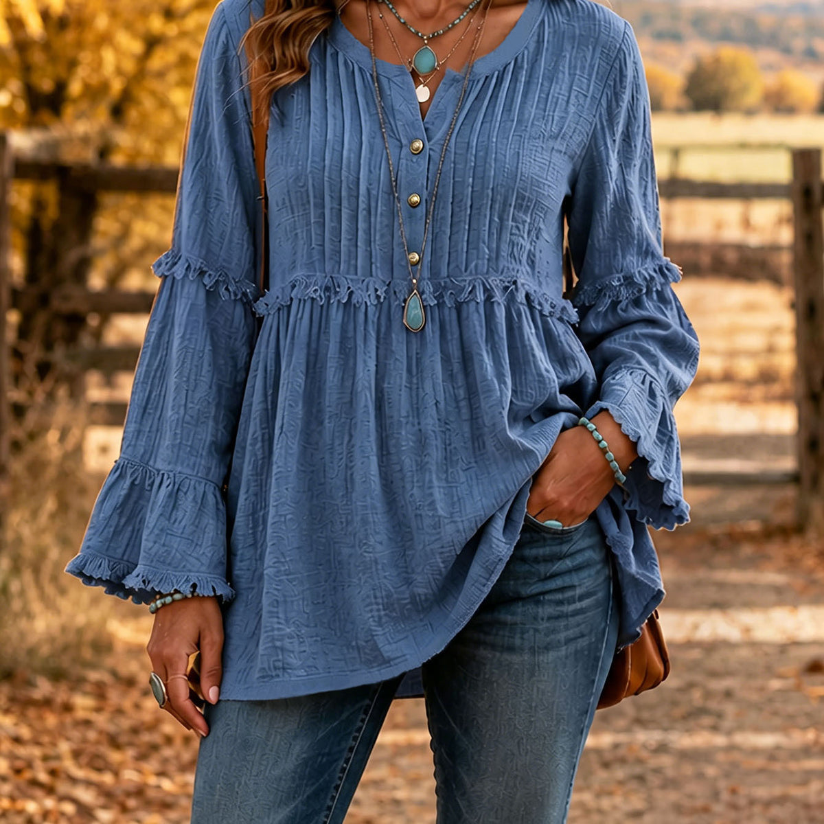 A woman wearing a blue long-sleeve blouse with ruffled trim and a button placket, paired with blue jeans, standing outdoors in front of a rustic wooden fence.