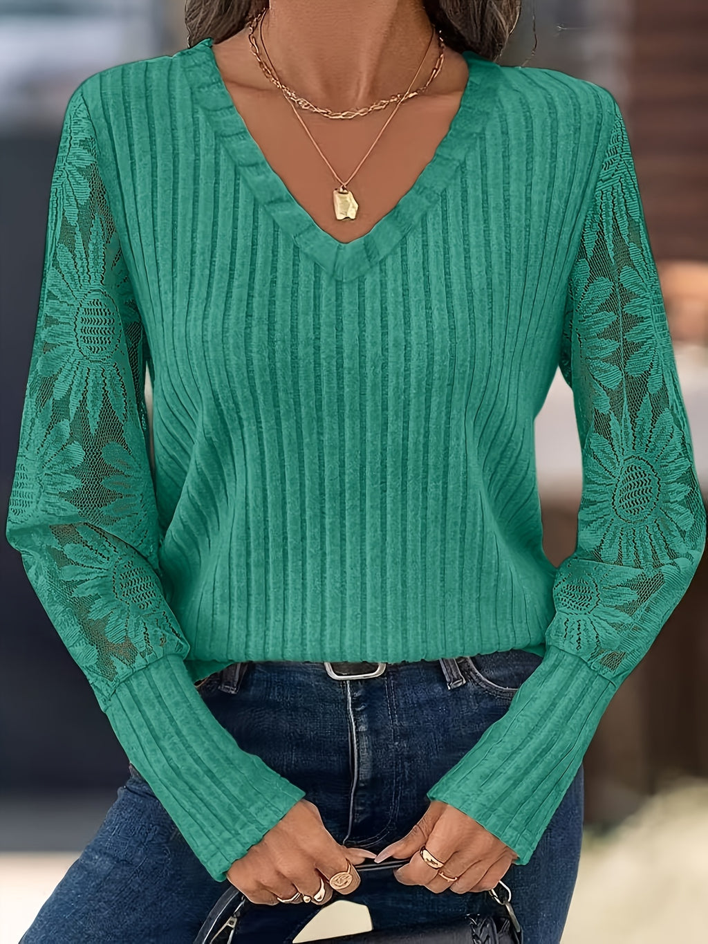 A woman wearing a green ribbed knit long-sleeve sweater with sheer sunflower-patterned sleeves, paired with blue jeans and layered gold necklaces.