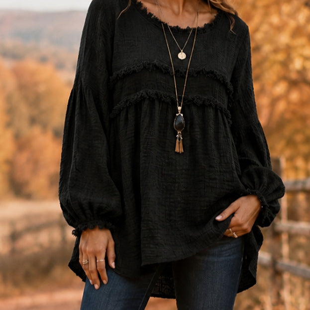 A woman wearing a black long-sleeve blouse with layered ruffle trim and a loose, flowy silhouette, paired with blue jeans and layered necklaces, set against an outdoor autumn background.