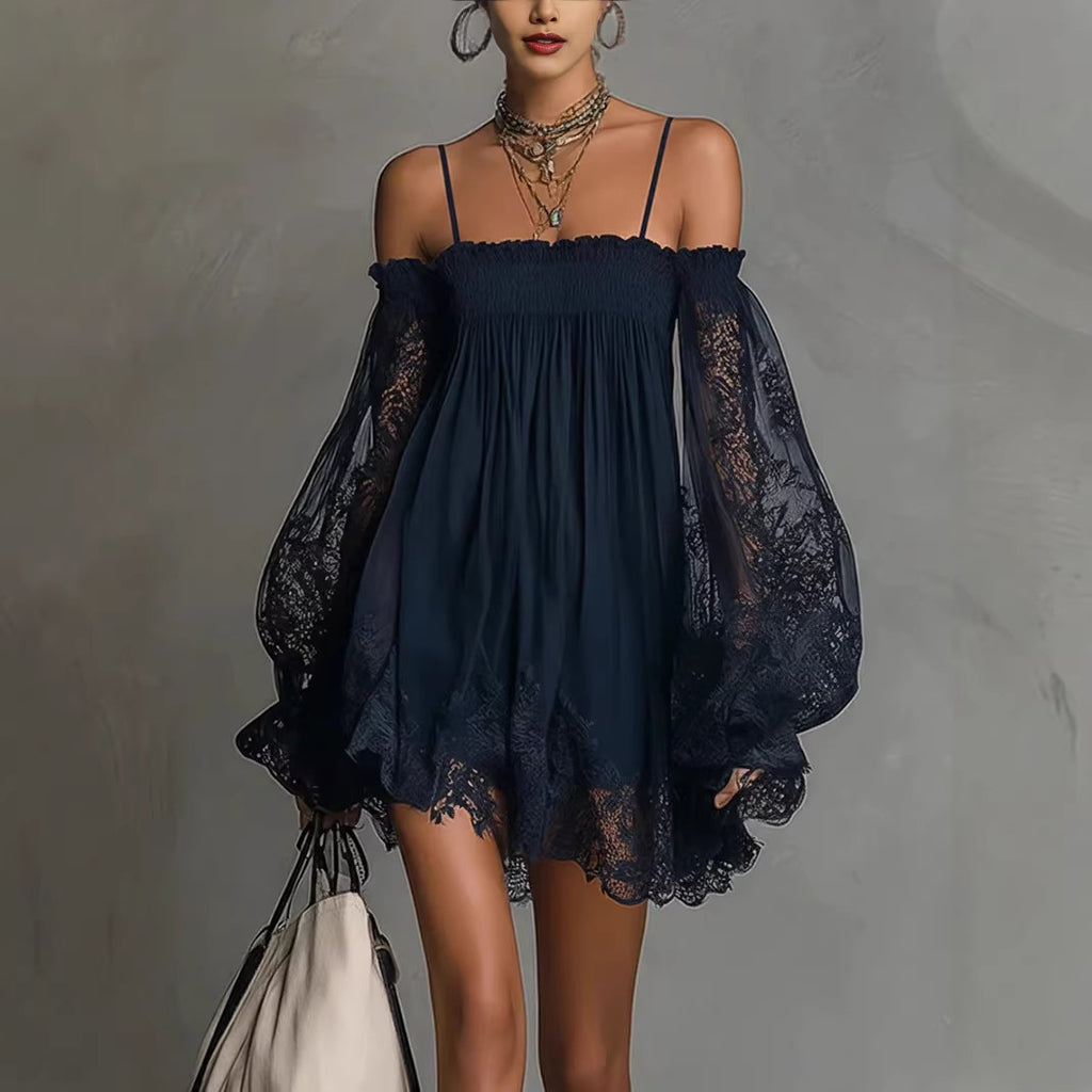 A woman models a short off-the-shoulder navy blue dress with sheer lace balloon sleeves and a smocked bodice, styled with layered necklaces and a cream shoulder bag against a textured gray wall.