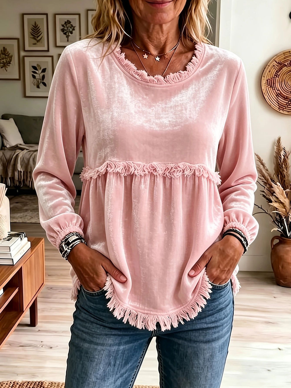 A woman wearing a light pink velvet long-sleeve top with ruffle trim and fringe details, paired with blue jeans, standing in a living room setting.