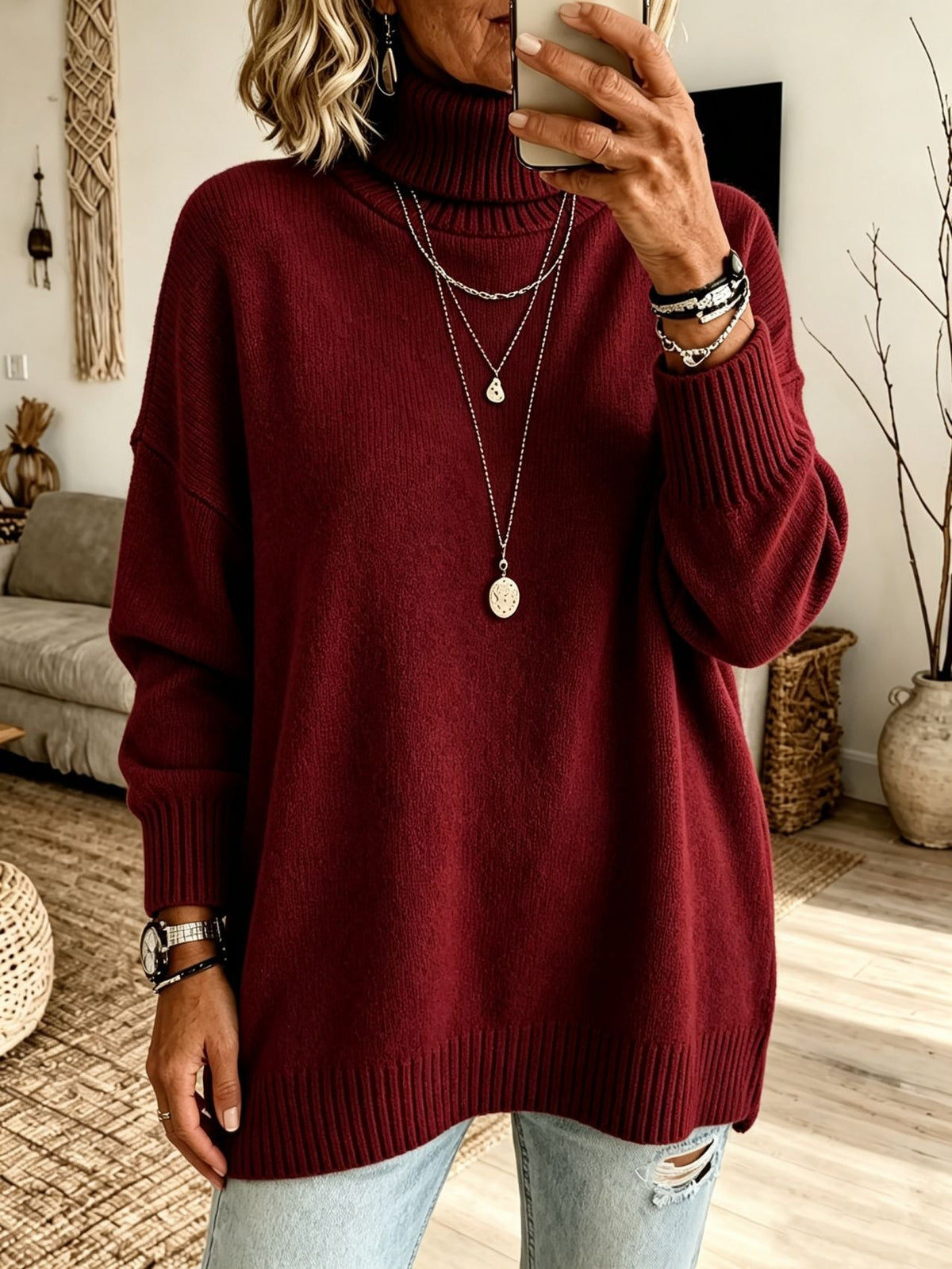 A woman wearing an oversized ribbed turtleneck sweater in deep burgundy, paired with light-wash distressed jeans and layered silver necklaces, taking a mirror selfie in a cozy living room setting.