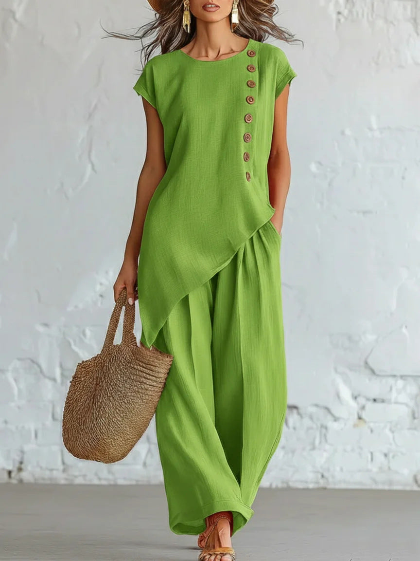 Woman wearing a green outfit with wide-leg pants and a straw bag against a white textured wall.