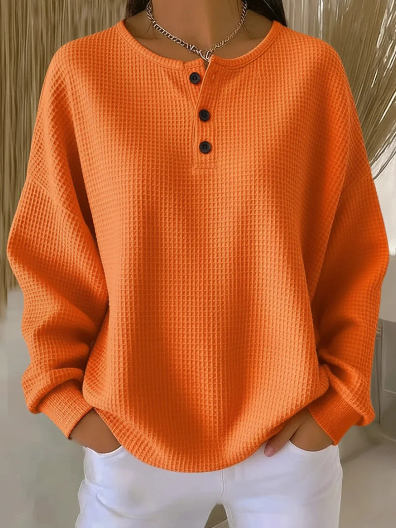 A person wearing a bright orange waffle-knit long-sleeve henley top with three dark buttons, paired with white pants, shown from the neck down in an indoor setting.
