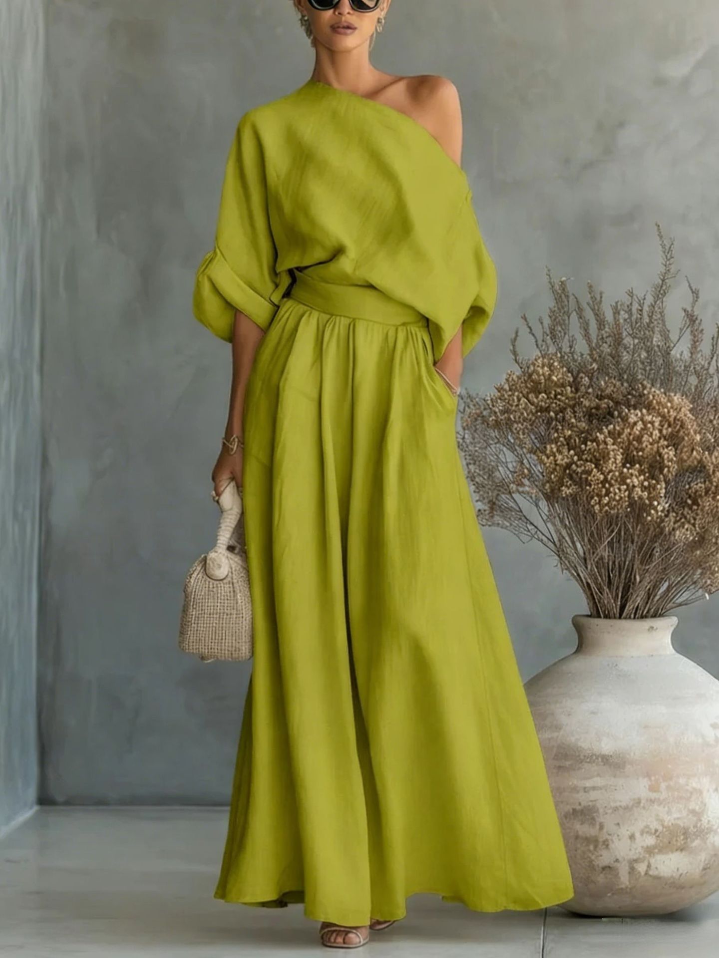 Woman wearing a green one-shoulder dress standing against a gray wall with a vase and dried plants.