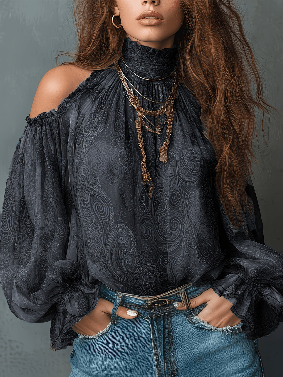 A close-up of a model wearing a dark charcoal gray cold-shoulder blouse with a high mock neck and a subtle paisley pattern, paired with frayed blue jeans and layered gold necklaces.