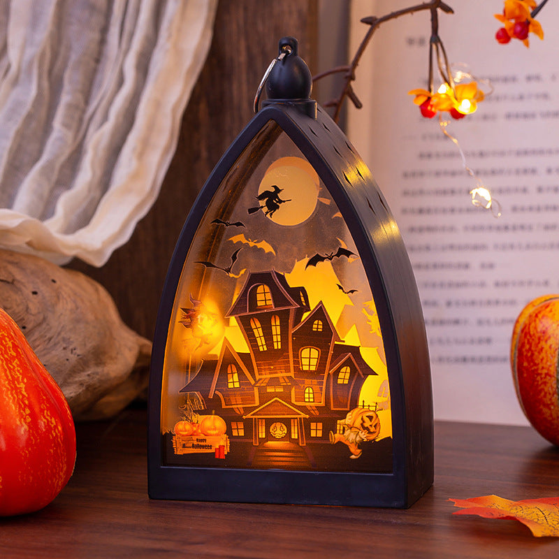 A Halloween-themed decorative lantern with a dark frame and a glowing cutout scene of a haunted house, bats, a witch on a broomstick, and a full moon, displayed on a tabletop with pumpkins and autumn decor in the background.