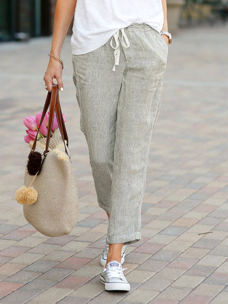 Person wearing light gray pants and white sneakers, holding a textured handbag with pom-poms on a paved walkway.