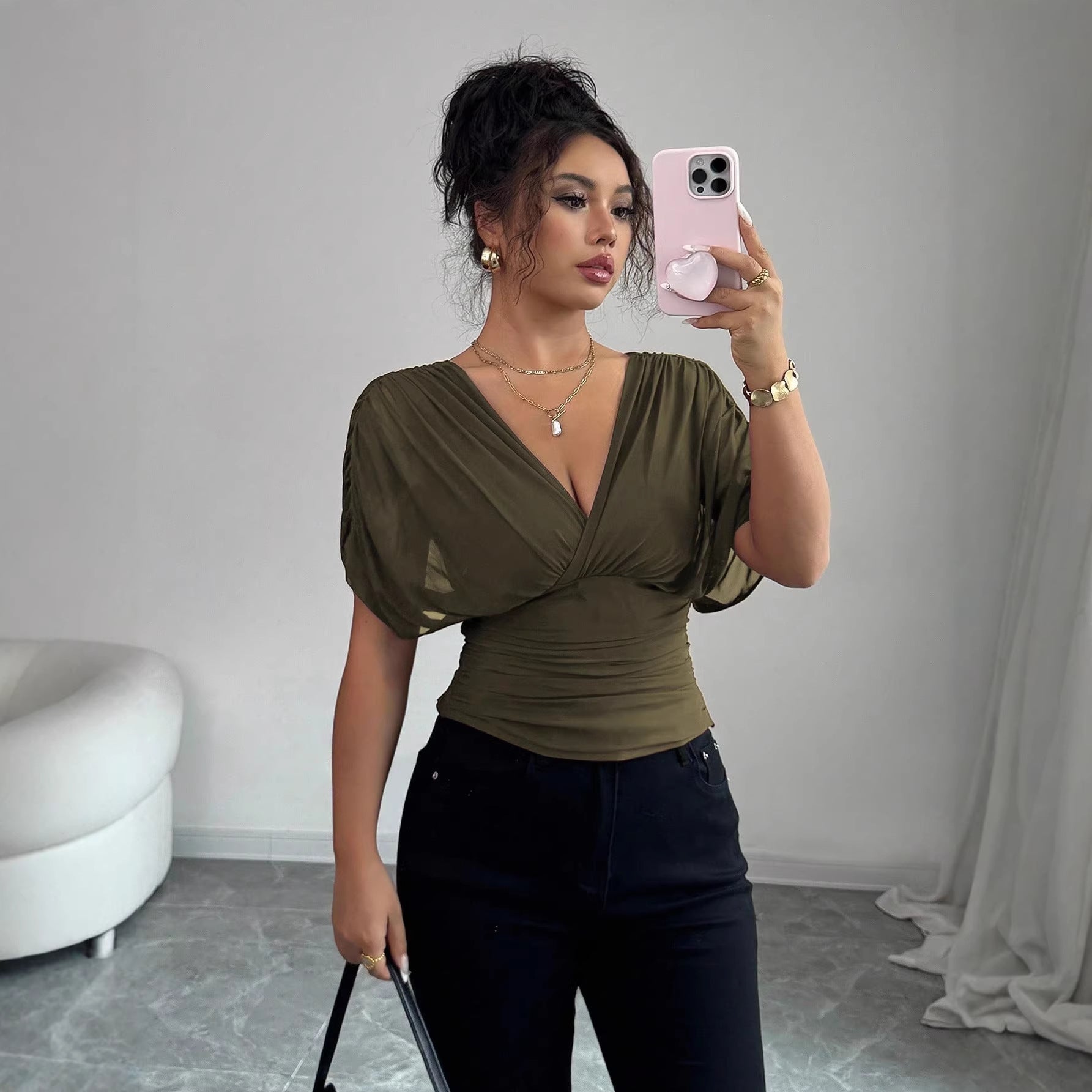 Woman taking a mirror selfie wearing an olive green top and black pants in a room with a white couch and gray floor.