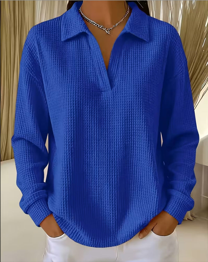 A woman wearing a bright royal blue waffle-knit long-sleeve collared shirt with a split V-neck, paired with white pants and a silver necklace.