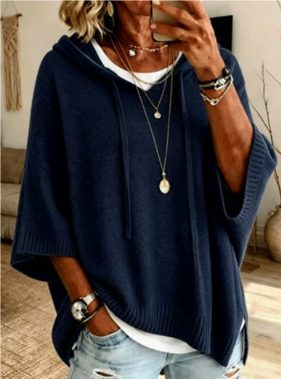 A woman wearing a navy blue knit poncho hoodie over a white t-shirt and light-wash distressed jeans, styled with layered gold necklaces and bracelets in an indoor setting.
