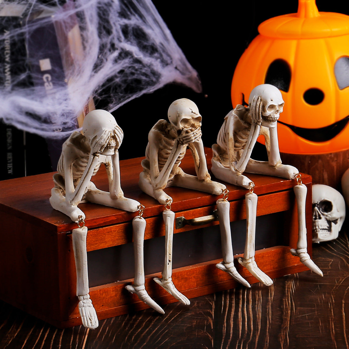 Three skeleton figurines on a wooden box with a Halloween pumpkin in the background.