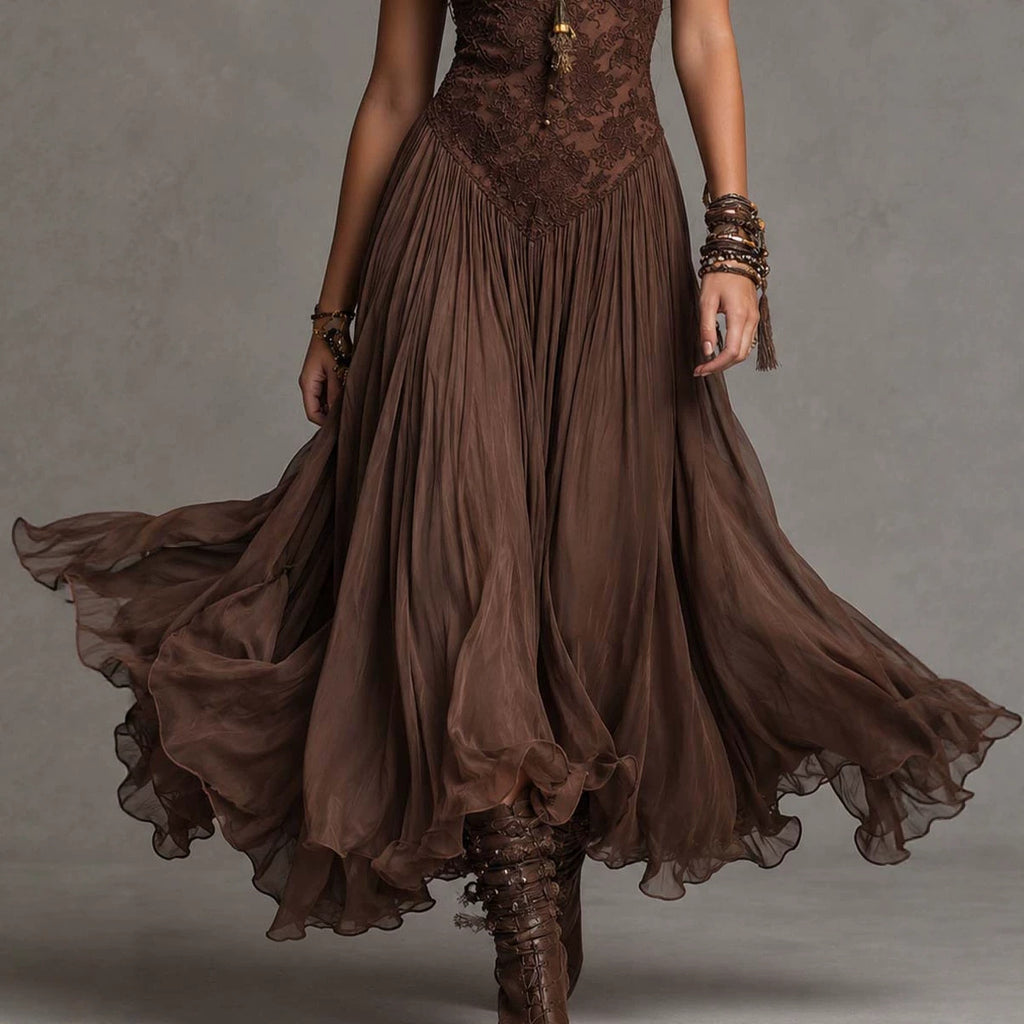 A woman wearing a flowing brown bohemian-style maxi dress with a lace bodice and sheer layered skirt, captured mid-motion against a neutral gray studio background.