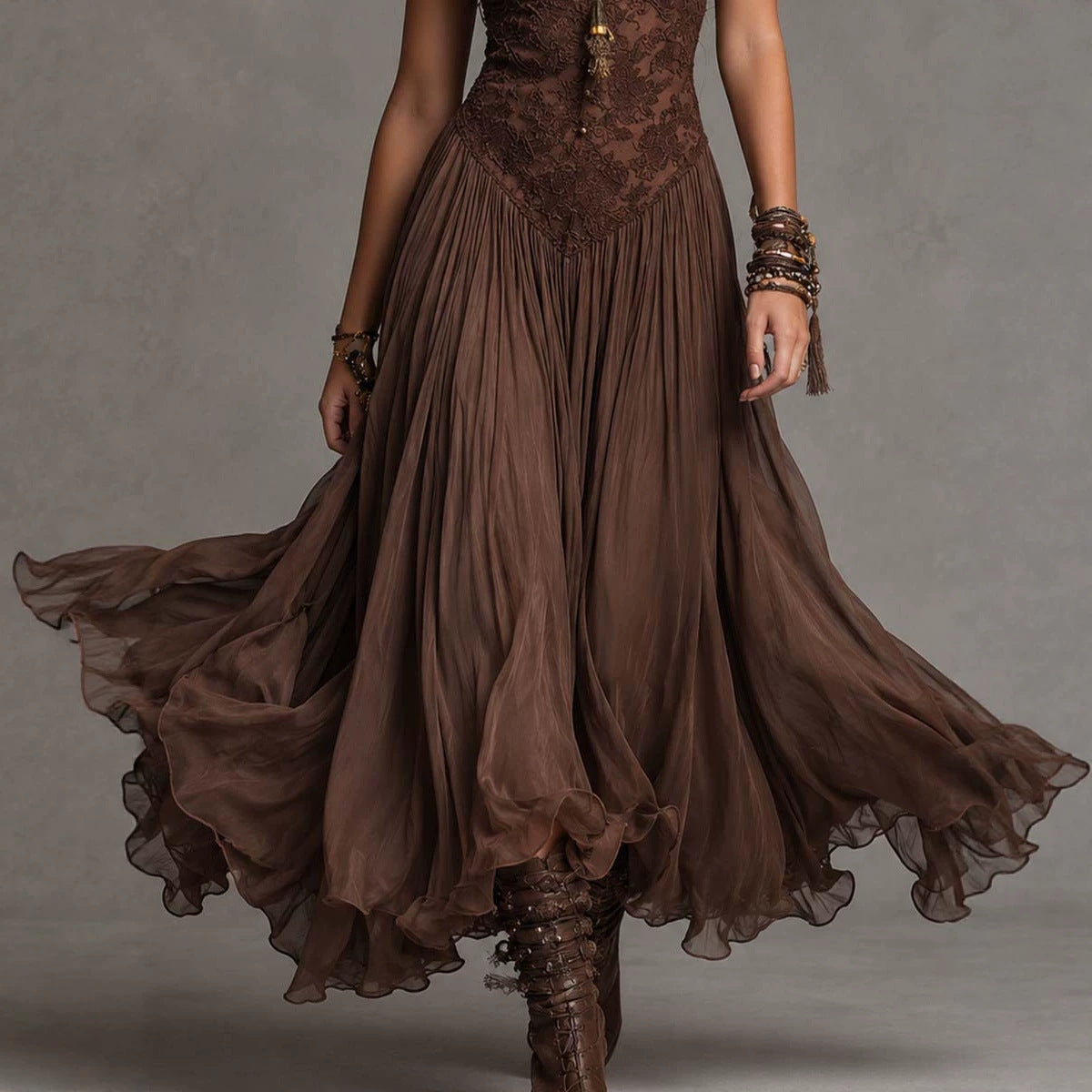 A woman wearing a flowing brown bohemian-style maxi dress with a lace bodice and sheer layered skirt, captured mid-motion against a neutral gray studio background.