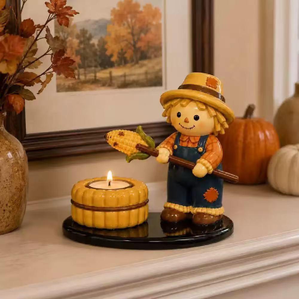 A decorative scarecrow figurine holding a corn cob stands beside a small yellow candle holder on a black base, displayed on a mantel with pumpkins, a framed autumn landscape, and dried leaves in the background.