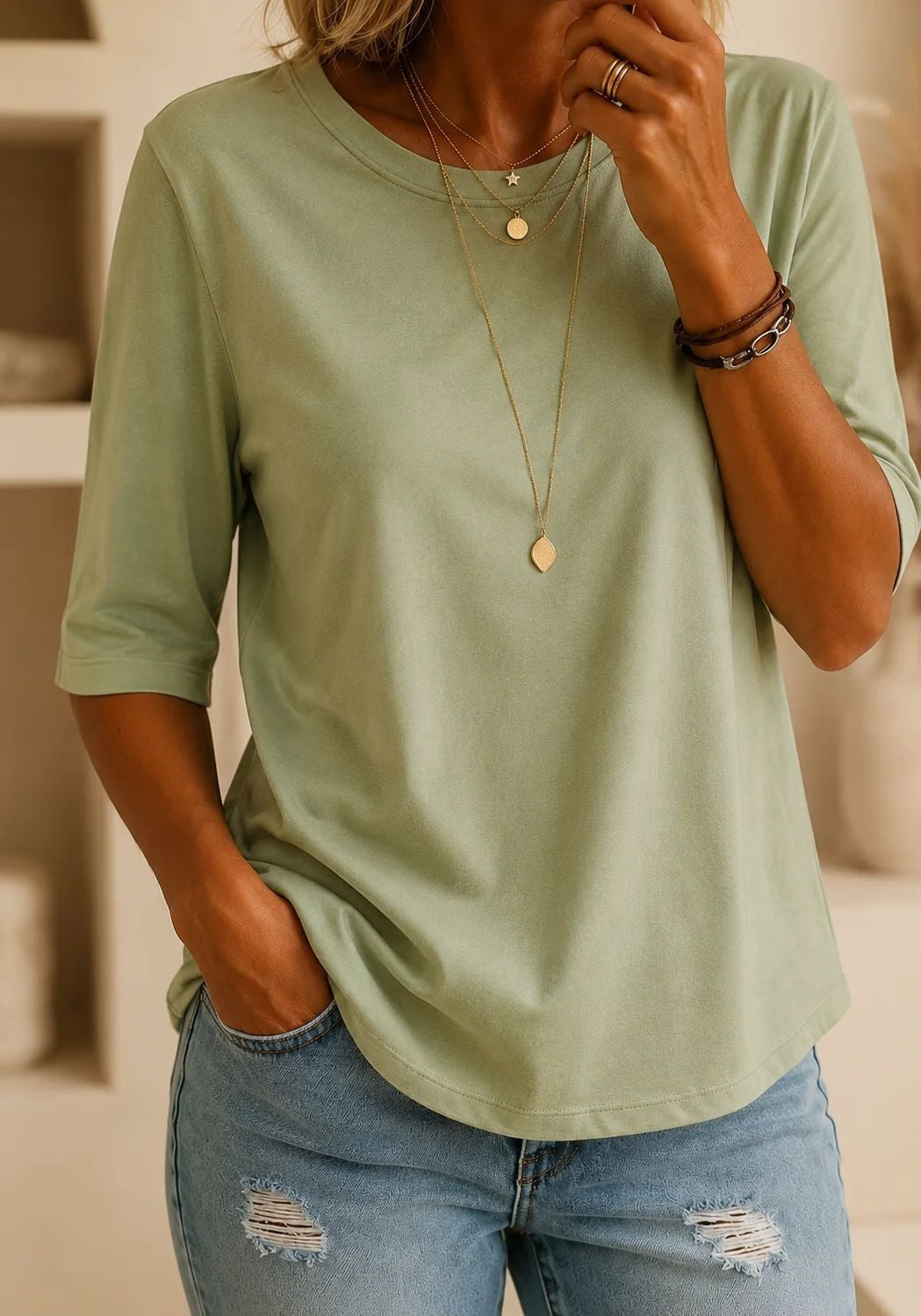 A woman wearing a sage green short-sleeve crewneck t-shirt and distressed blue jeans, accessorized with layered gold necklaces and bracelets.