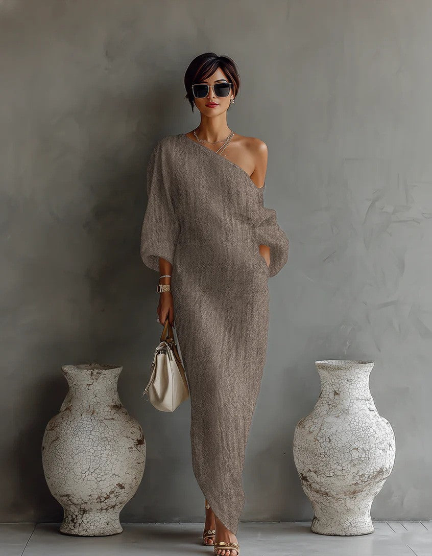 Woman in a beige one-shoulder dress standing between two large vases against a gray wall.