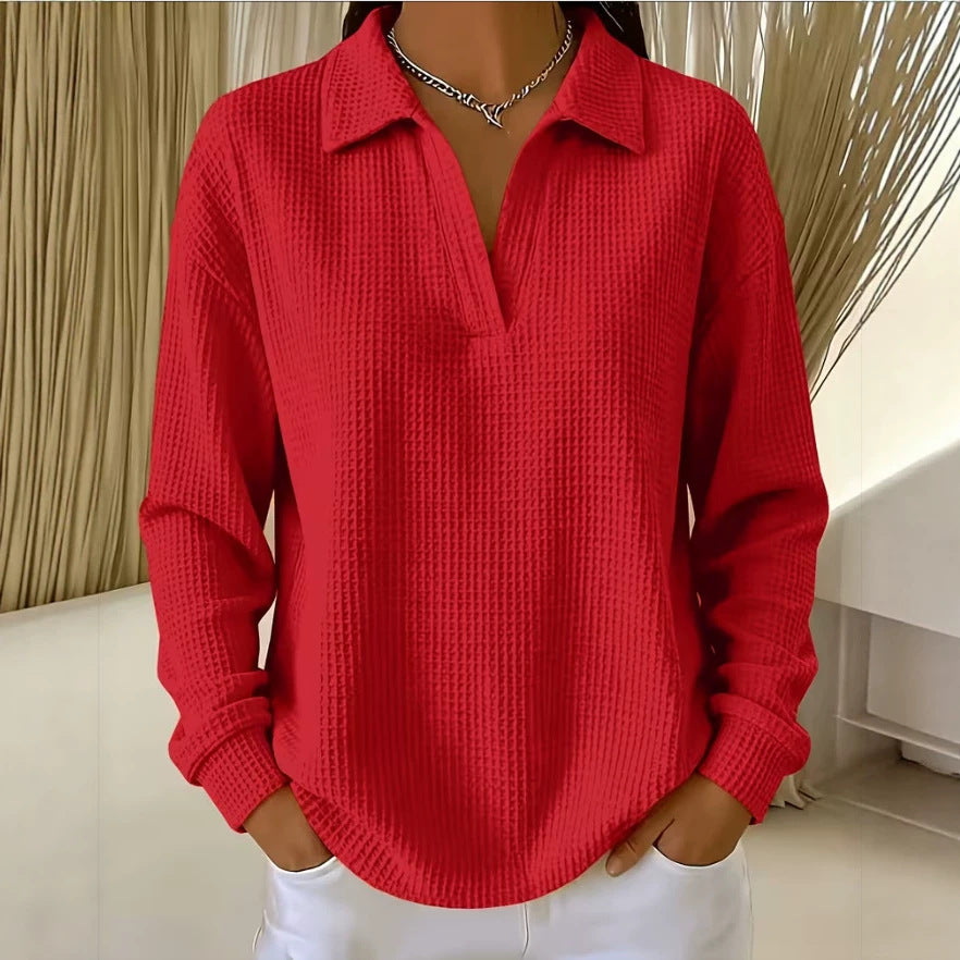 A woman wearing a bright red waffle-knit long-sleeve polo shirt with a collar and split neckline, paired with white pants, standing indoors.