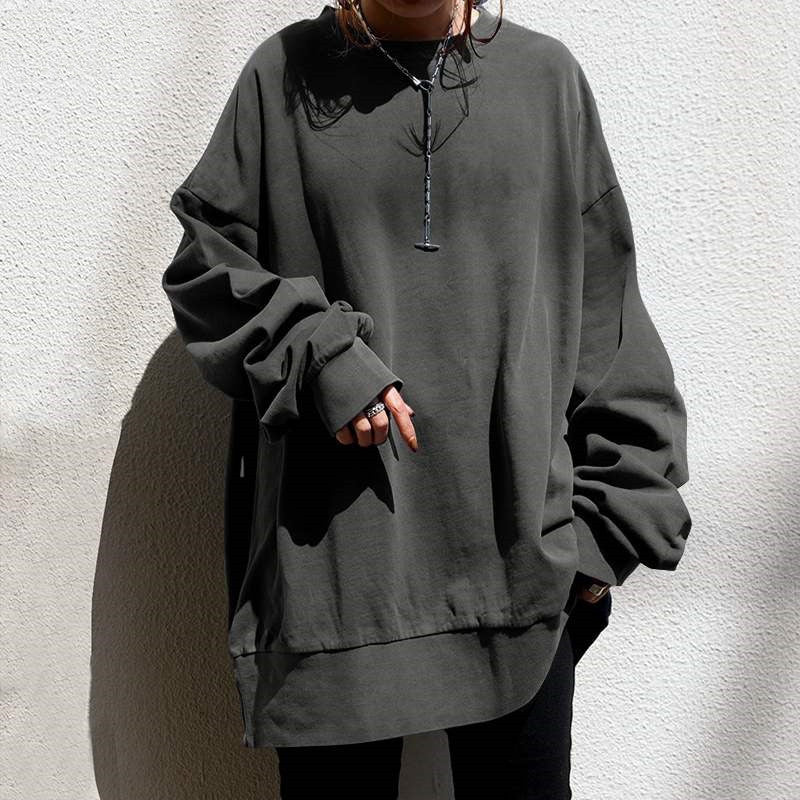 A person wearing an oversized dark charcoal gray sweatshirt with a half-zip neckline, paired with black pants, standing against a textured off-white wall.