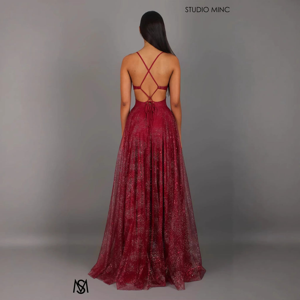 Woman wearing a burgundy evening gown with a criss-cross back design on a gray background, with 'Studio Minc' branding.