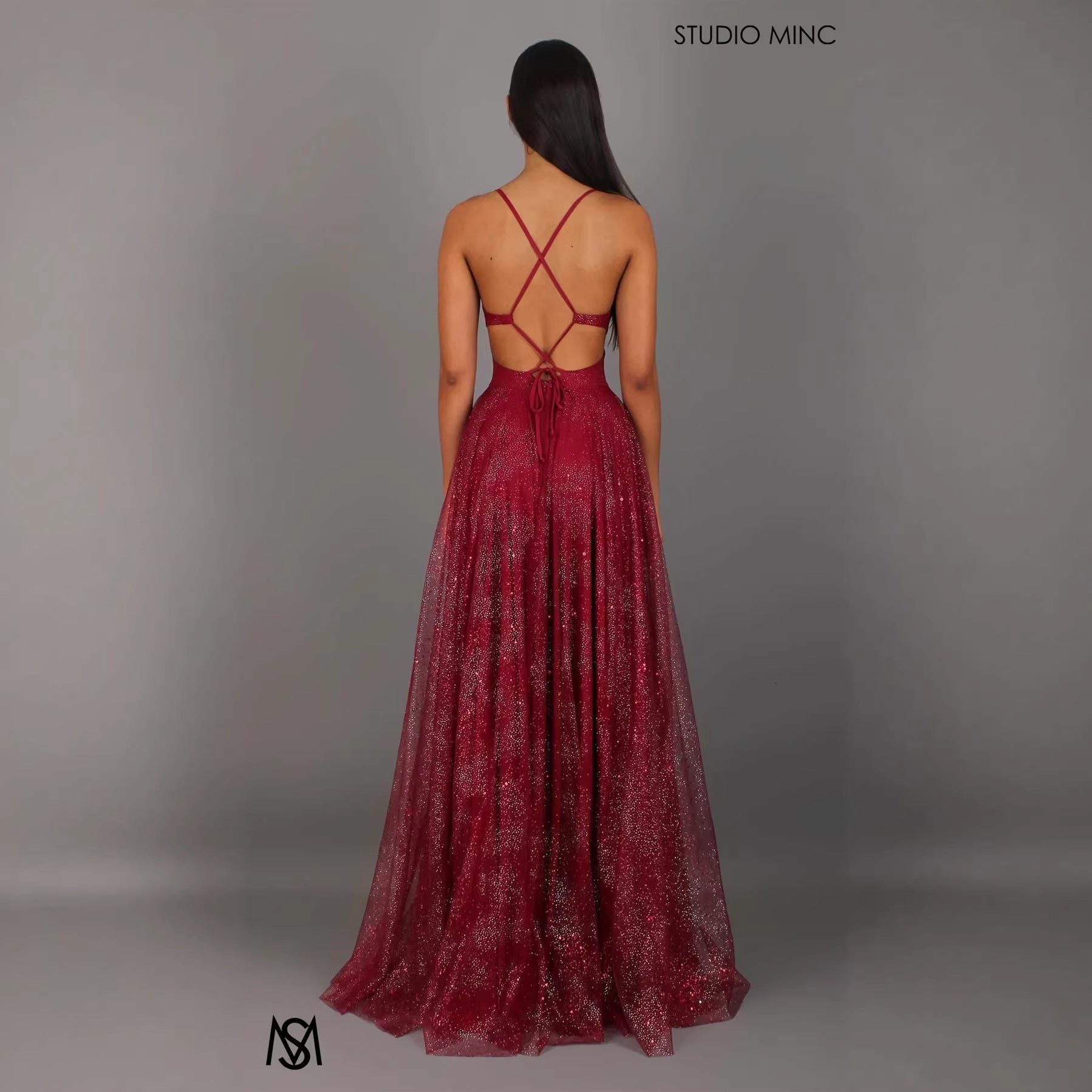 Woman wearing a burgundy evening gown with a criss-cross back design on a gray background, with 'Studio Minc' branding.
