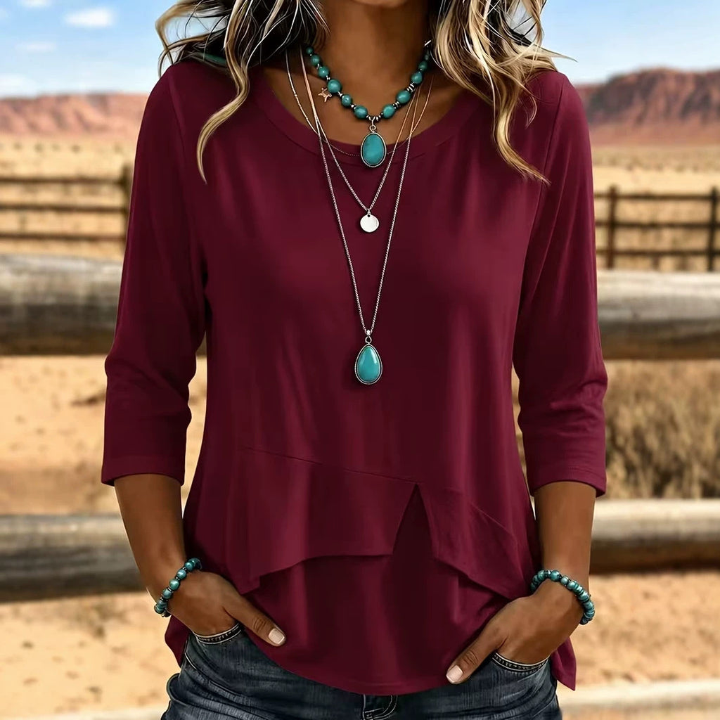 A woman wearing a three-quarter sleeve burgundy top with a layered ruffle hem, styled with turquoise jewelry, standing in a desert landscape.