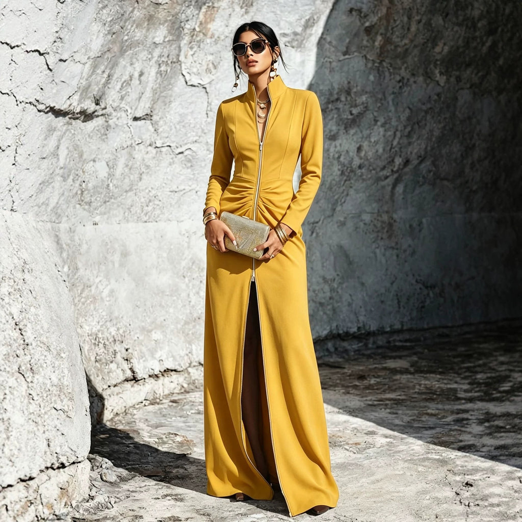 A woman wearing a long-sleeve mustard yellow maxi dress with a front zipper and high slit, holding a metallic clutch against a textured stone wall.