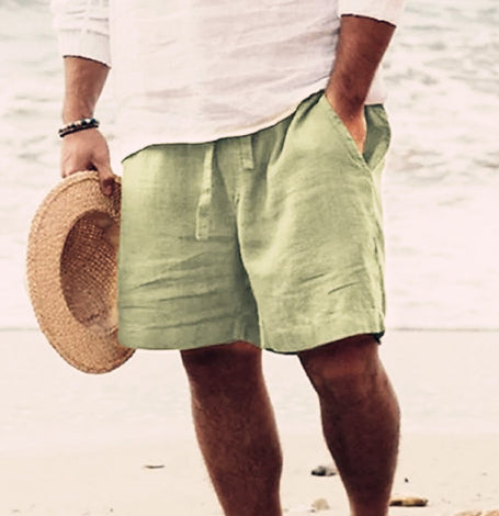 Person wearing green shorts and holding a straw hat on a beach.