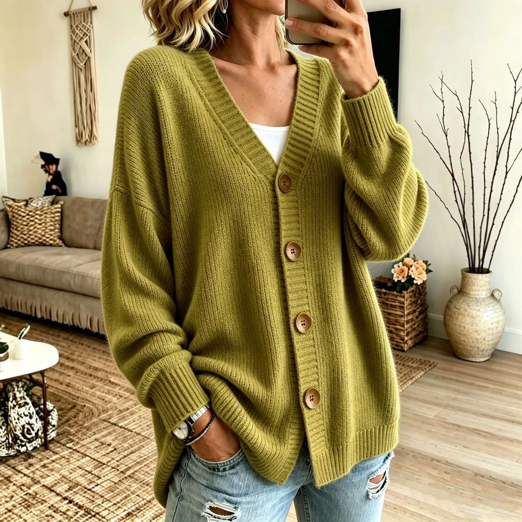 A woman wearing an olive green ribbed knit cardigan with brown buttons, styled with light blue distressed jeans in a cozy living room setting.