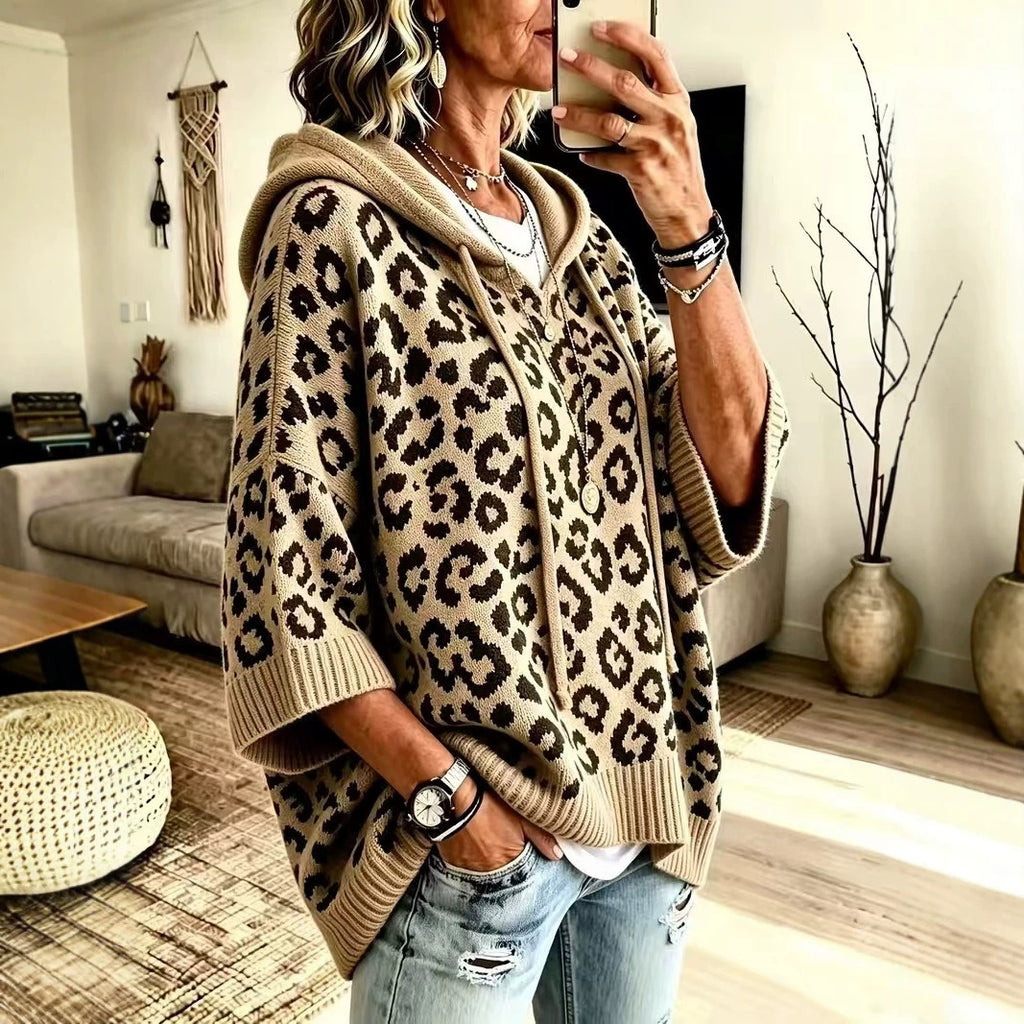 A woman takes a mirror selfie in a cozy living room while wearing a beige and brown leopard-print knit hoodie poncho sweater with ribbed cuffs and hem.