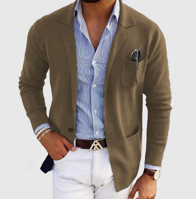 Man wearing a green blazer over a blue striped shirt with white pants on a light gray background