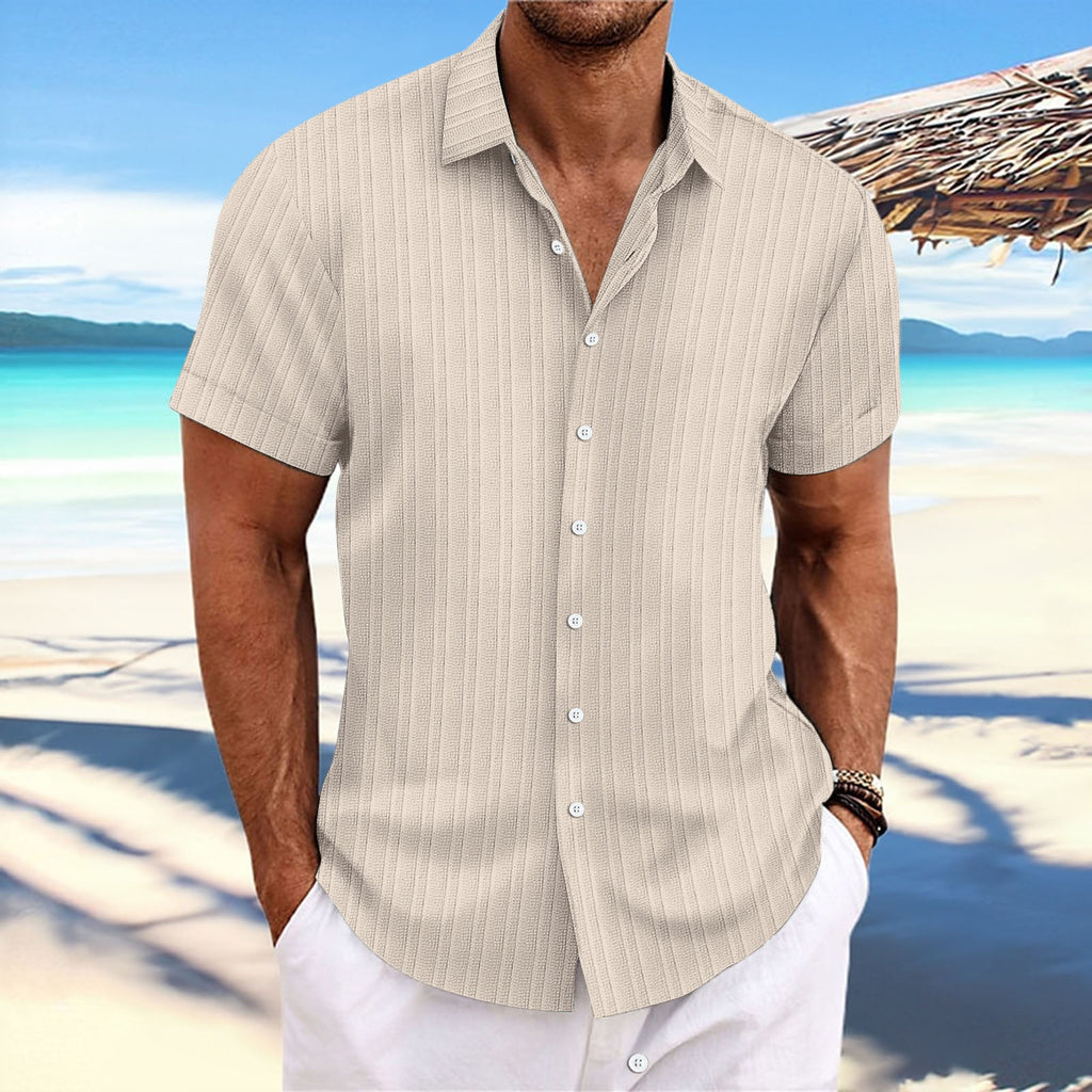 Man wearing a beige striped shirt and white pants on a beach with clear blue water and sky.
