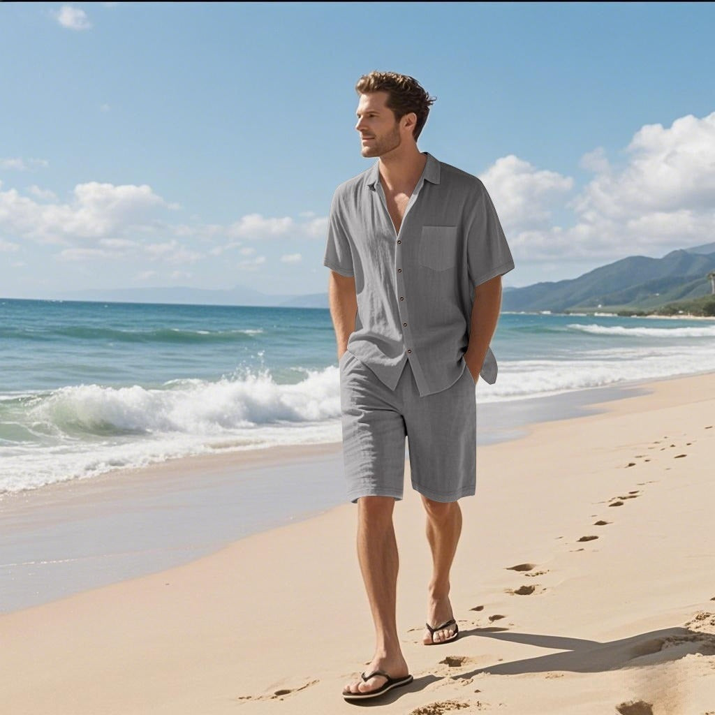 Man in gray shirt and shorts standing on a sandy beach with ocean and mountains in the background