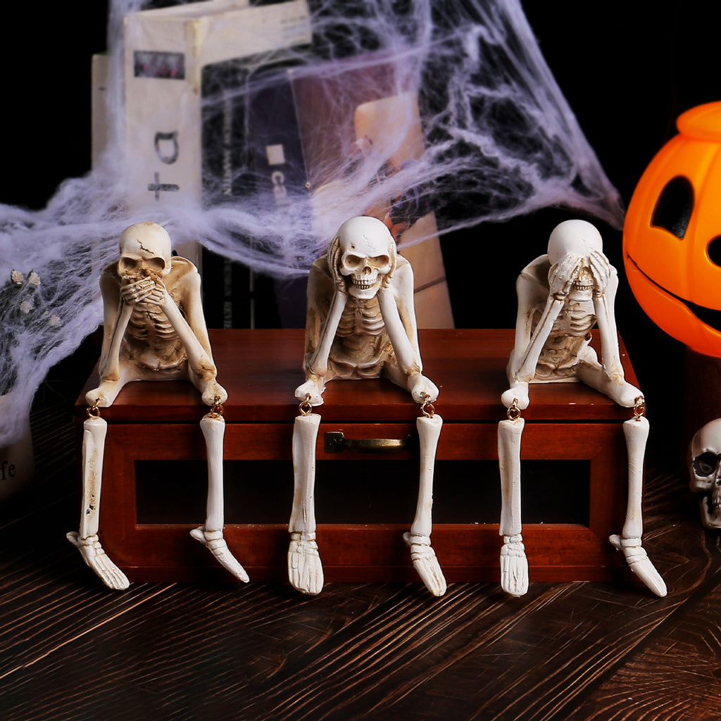 Three skeleton figurines on a wooden box with Halloween decorations in the background.