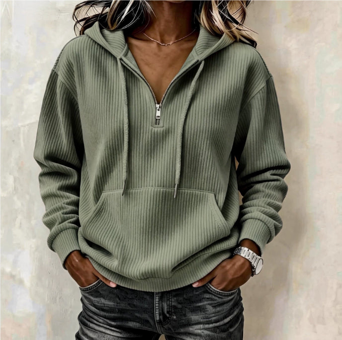 A woman wearing an olive green ribbed pullover hoodie with a half-zip neckline, drawstrings, and a front kangaroo pocket, paired with dark jeans against a textured beige wall.