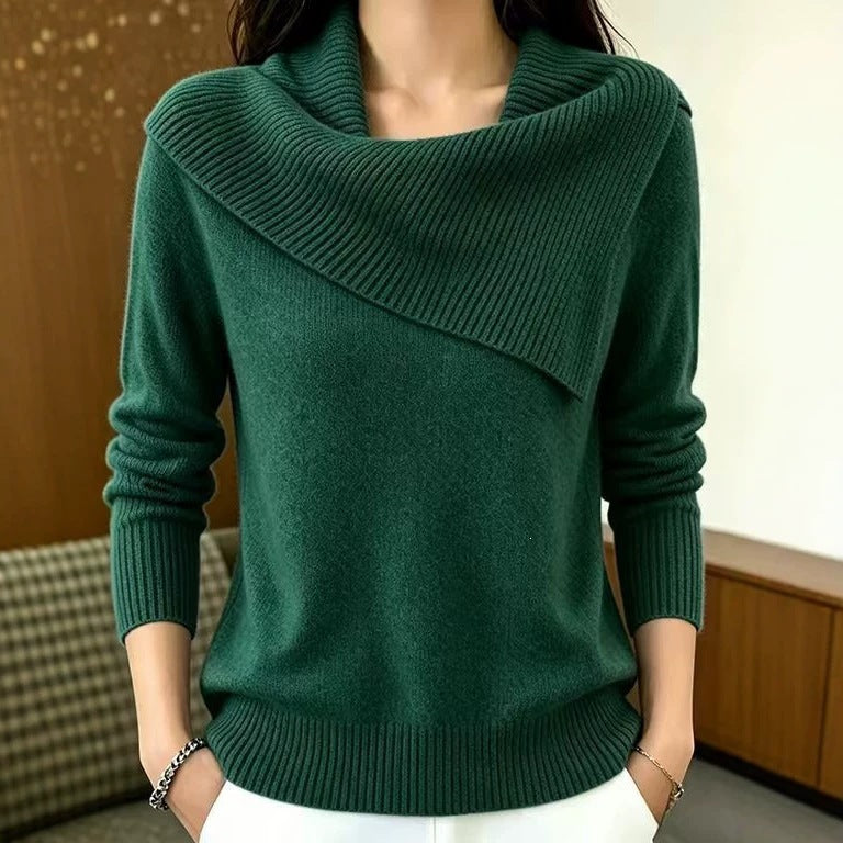 A woman wearing a dark green knit sweater with a wide draped cowl neckline, long sleeves, and ribbed trim, paired with white pants in an indoor setting.
