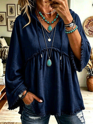 A woman wearing a navy blue bohemian-style tunic top with fringed edges, layered necklaces, and turquoise jewelry, paired with distressed jeans in an indoor setting.