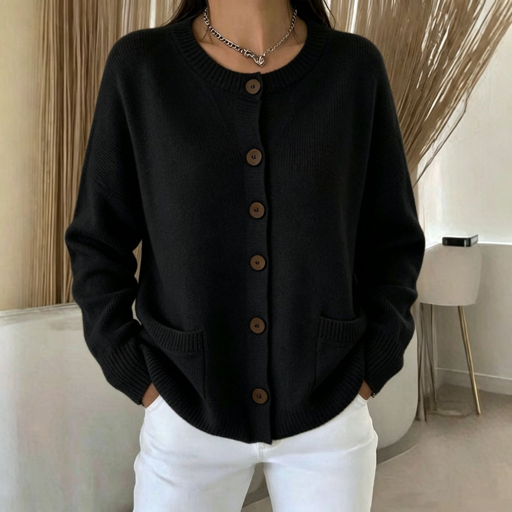 A person wearing a black knit cardigan sweater with brown buttons and white pants, standing indoors near a chair and dried decorative branches.