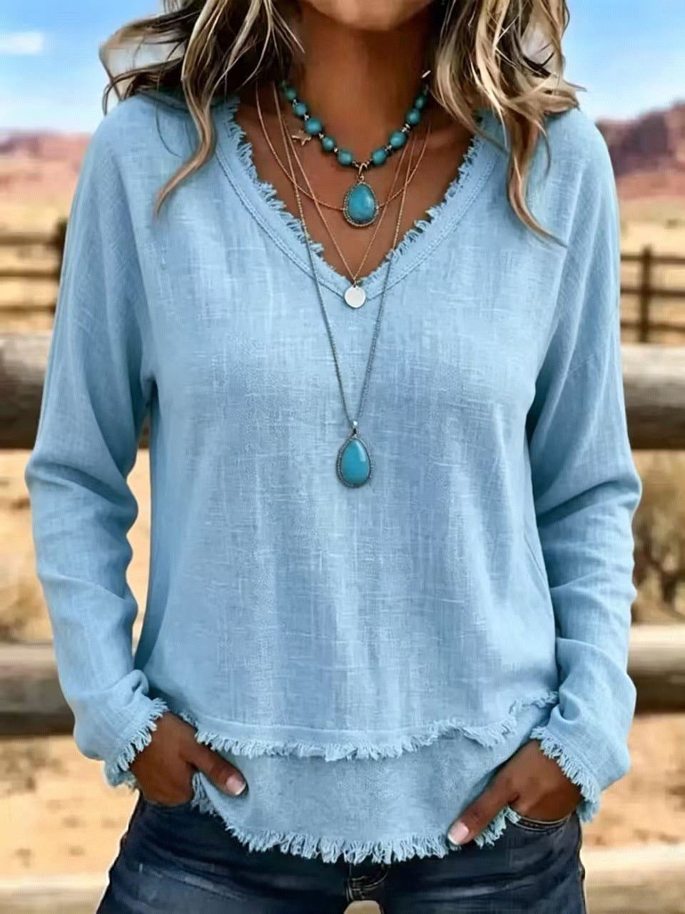 A woman wearing a light blue long-sleeve top with a frayed hem and neckline, layered with turquoise and silver necklaces, standing outdoors in a desert landscape.