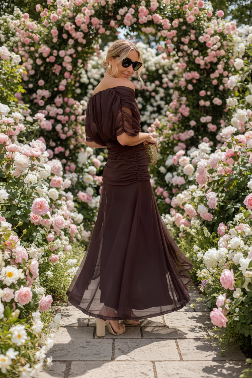 Woman wearing a dark brown off-shoulder dress with sheer sleeves background of flowers and pavement.