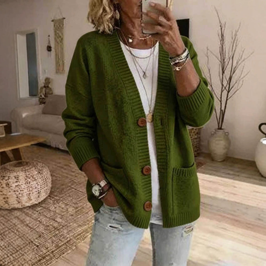 A woman takes a mirror selfie wearing an olive green knit cardigan with brown buttons over a white t-shirt and light-wash distressed jeans, in a cozy living room setting.