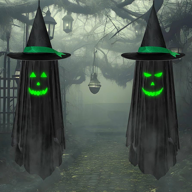 Two black witch ghost decorations with glowing green jack-o'-lantern faces hang on either side of a foggy, spooky forest path.
