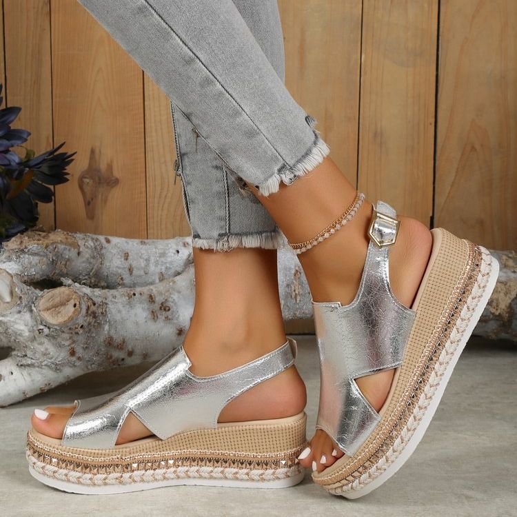 Silver sandals with woven straps worn by a person on a wooden floor.