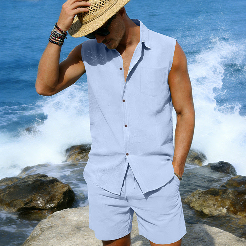Man in light blue sleeveless shirt and shorts standing by the ocean with waves crashing around.
