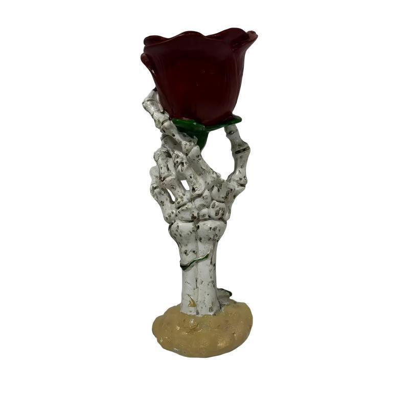 A skeleton hand figurine holding a dark red rose with green leaves, mounted on a beige base against a plain white background.
