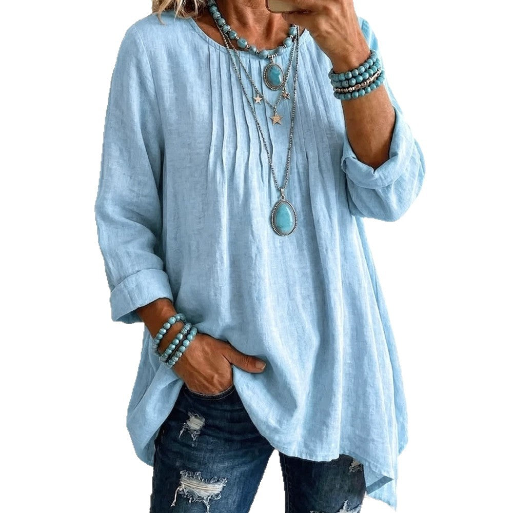 A woman wearing a light blue linen-textured long-sleeve tunic top with a gathered neckline and pockets, paired with distressed blue jeans and layered turquoise jewelry, shown in a mirror selfie style against a white background.