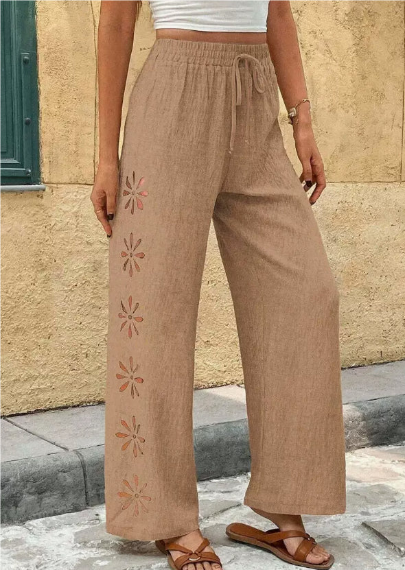 Person wearing brown linen pants with floral embroidery against a stone wall.