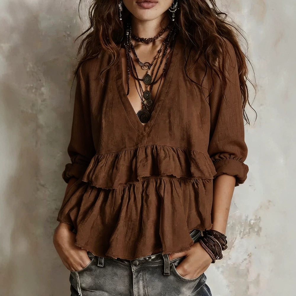 A woman wearing a brown long-sleeve bohemian-style blouse with a tiered ruffle hem, layered with multiple dark necklaces and bracelets, paired with gray jeans against a textured neutral wall.