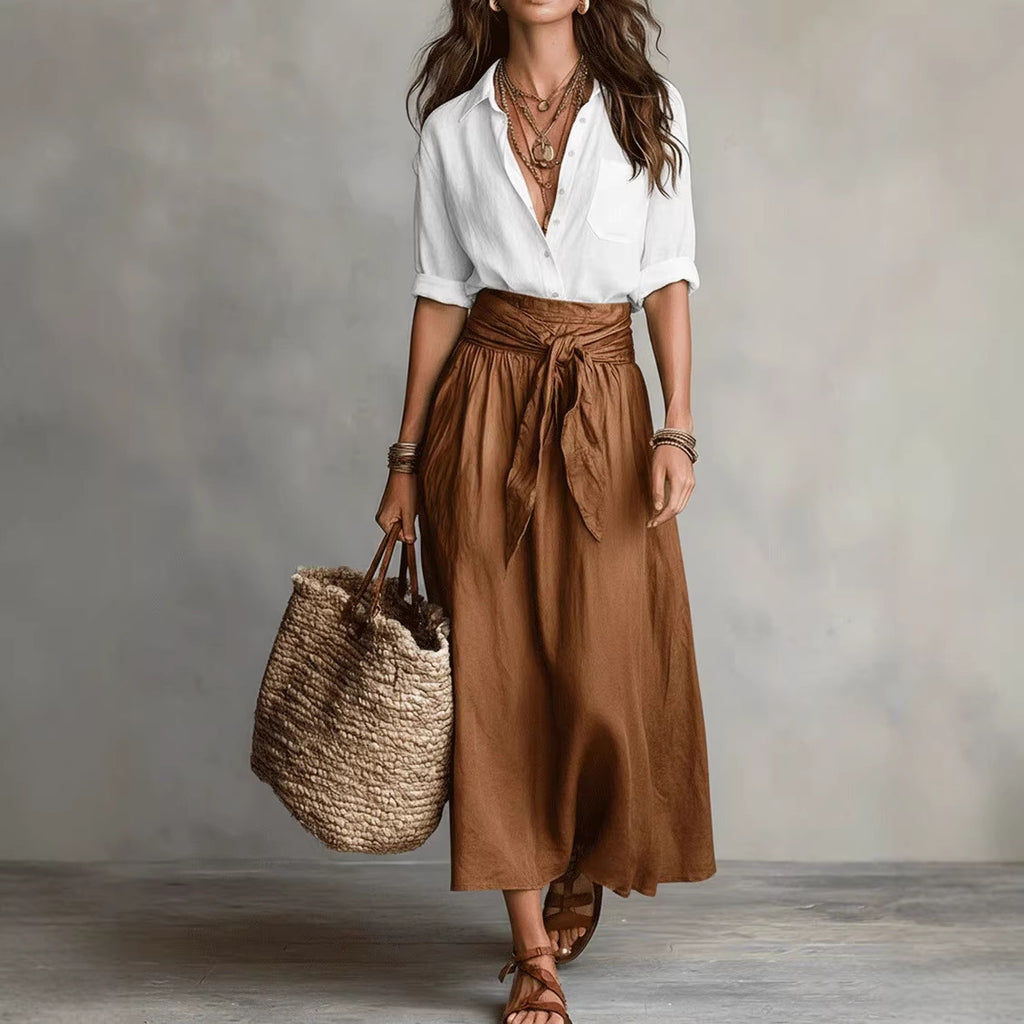 Woman wearing a white shirt and brown skirt holding a woven bag against a neutral background