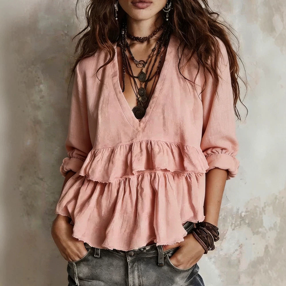 A woman wearing a dusty pink bohemian-style blouse with a deep V-neck, ruffled tiers, and rolled sleeves, paired with layered dark brown necklaces and dark gray jeans, set against a textured gray wall.